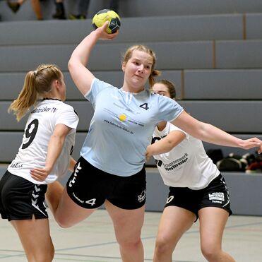 Handballspielerin in blauer Trikot wirft den Ball, während zwei gegnerische Spielerinnen sie versuchen zu blockieren.