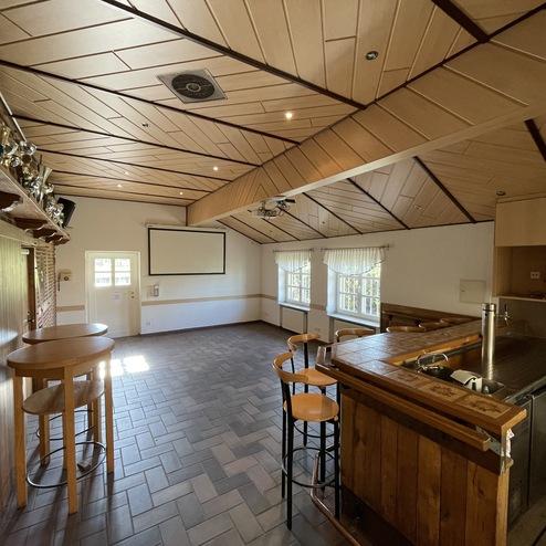 Leerer, lichtdurchfluteter Raum mit Theke, Hockern, und einer Leinwand an der Wand. Wandregal mit Pokalen.