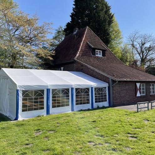Weißes Zelt mit blauen Akzenten steht vor einem roten Backsteinhaus umgeben von grünem Rasen und Bäumen.