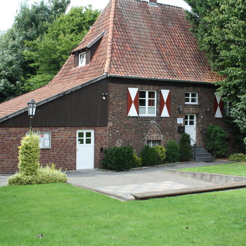 Historisches Haus mit rotem Dach und roten Fensterläden, umgeben von Rasen und Büschen in einem grünen Garten.