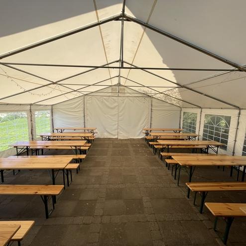 Blick in ein großes Zelt mit mehreren Holztischen und Bänken, beleuchtet durch Tageslicht und mit Fensterfront.