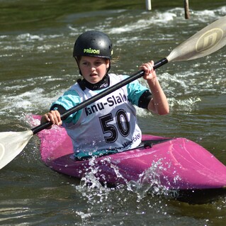 Junge im pinken Kajak paddelt konzentriert durch eine bewegte Wasserstrecke während eines Wettkampfs.