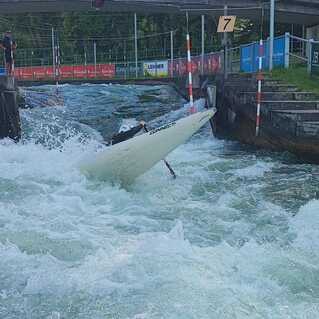 Kajakfahrer steuert durch schäumende Wellen, während ein Zuschauer auf einer Plattform zusieht.
