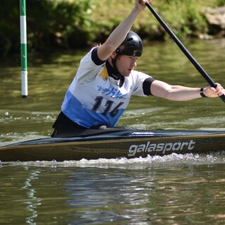 Ein Kanute paddelt intensiv in einem Fluss, mit einer grünen Tore markierung im Hintergrund.