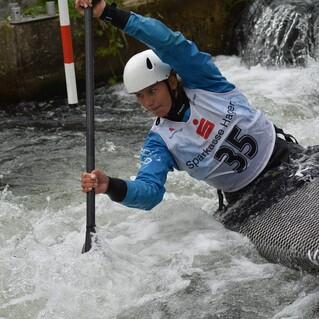 Sportlerin paddelt energisch in einem Wildwasser-Kanu, umgeben von sprudelndem Wasser und einem Hindernis.