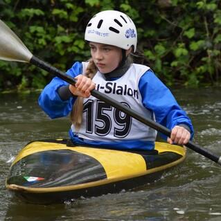 Mädchen im Kanu paddelt durch einen Fluss, trägt einen weißen Helm und ein sportliches Oberteil mit der Nummer 159.