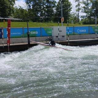 Kanufahrer manövriert durch strömendes Wasser in einem Wildwasserkanal, mit Toren am Ufer.
