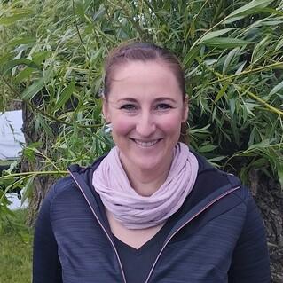 Lächelnde Frau mit leicht lockigem Haar, in schwarzer Jacke und pinkem Schal, vor einem Hintergrund aus Weidenzweigen.