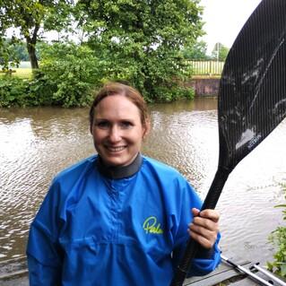 Eine Frau in blauer Paddeljacke lächelt und hält ein Paddel neben einem Wasserlauf mit Bäumen im Hintergrund.