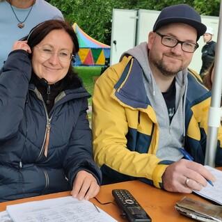 Zwei Personen sitzen an einem Tisch, eine spricht mit einem Headset, während die andere auf einem Blatt notiert.