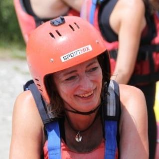 Lachende Frau in rotem Helm und Schwimmweste, bereit zum Rafting, mit bunten Booten im Hintergrund.
