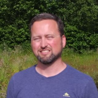 Mann in blauer T-Shirt steht in einer Wiese mit grünen Bäumen im Hintergrund, lächelnd und mit geschlossenen Augen.