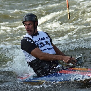 Sportler paddelt auf einem Kanuslalom-Kajak durch aufgewühltes Wasser, um Hindernisse zu umfahren.