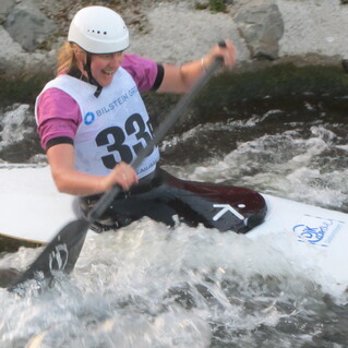 Eine Kanu-Fahrerin paddelt begeistert durch das rauschende Wasser, trägt einen Helm und eine Wettkampfnummer.