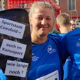 Drei Personen in blauen Shirts halten Paddel mit Aufschriften bei einer Veranstaltung in Coesfeld.