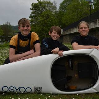 Drei Kinder in Neoprenanzügen posieren lächelnd vor einem Kajak auf einer Wiese mit blühenden Blumen.
