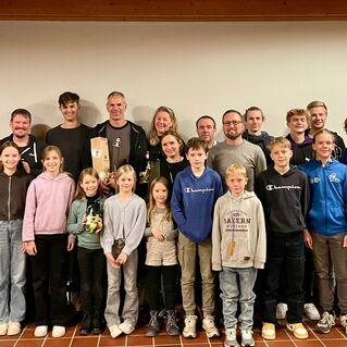 Gruppenfoto von Erwachsenen und Kindern in Sportkleidung, lächelnd vor einer neutralen Wand im Innenbereich.