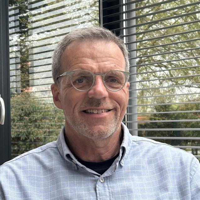 Mann mit Brille und grauen Haaren sitzt hinter einem Fenster mit horizontalen Jalousien und Blick ins Freie.
