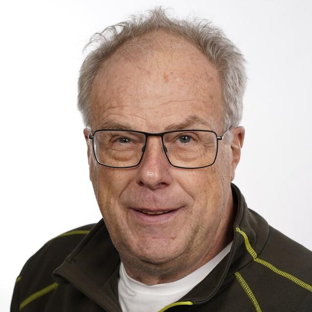 Porträt eines lächelnden Mannes mit Brille, grau melierten Haaren und einem dunkelgrünen Oberteil vor einem weißen Hintergrund.