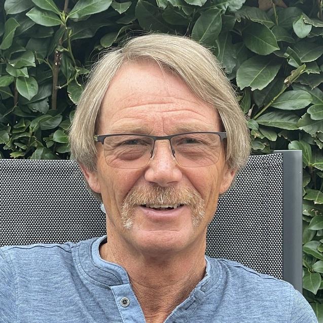 Mann mit Brille und einem Schnurrbart sitzt entspannt vor einer grünen Hecke, trägt ein blaues Langarmshirt.