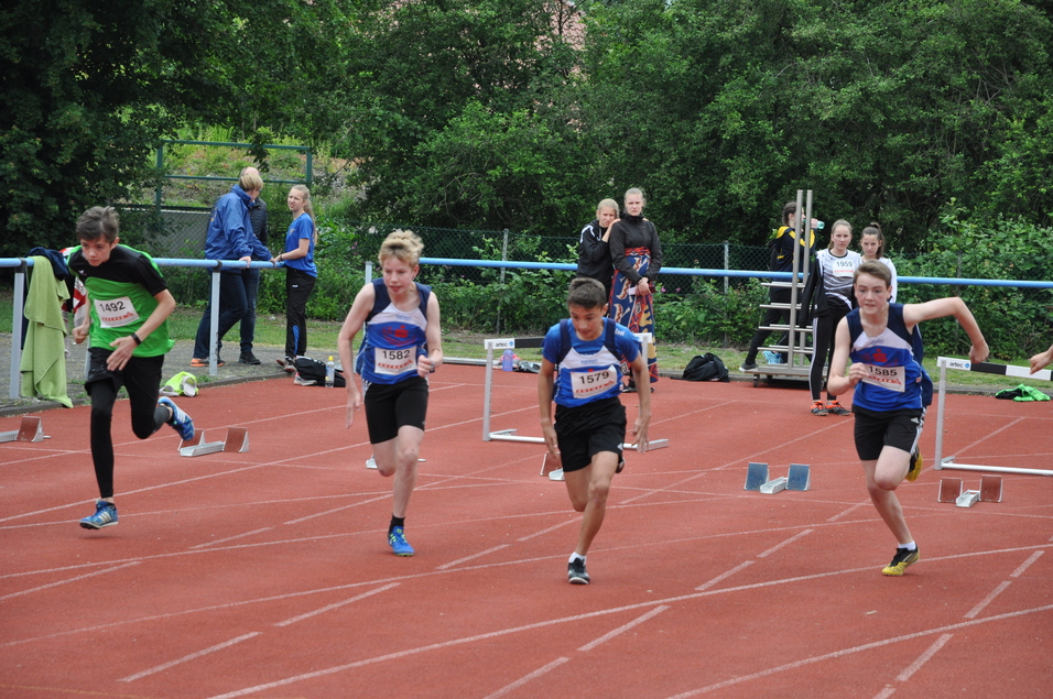Vier Schüler sprinten über eine rote Laufbahn bei einem Sportereignis, während Zuschauer im Hintergrund zuschauen.