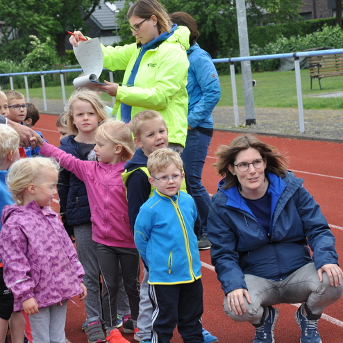 Grupp von Kindern steht auf einer Laufbahn, während eine Betreuerin Notizen macht und ein anderer Betreuer daneben steht.
