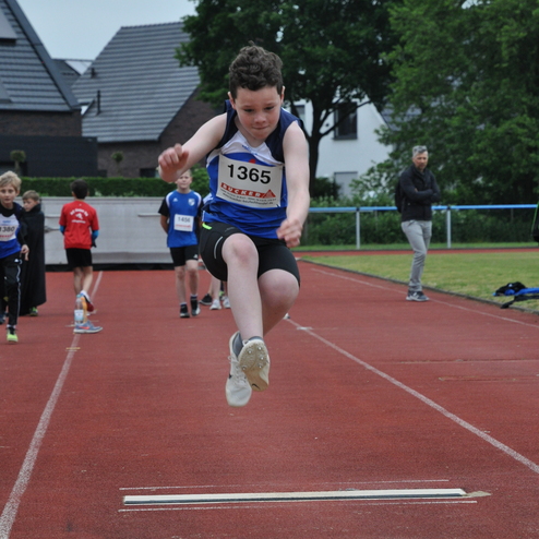 Ein Junge springt in einem Wettkampf von einer markierten Anlaufzone auf eine rote Laufbahn, im Hintergrund Zuschauer.