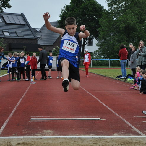 Ein Junge springt beim Weitsprung über die Grenze zur Sandgrube auf einer roten Sportbahn. Zuschauer sind im Hintergrund.