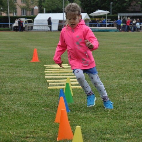 Mädchen in pinker Jacke überquert Hindernisse auf einem Sportplatz mit Kegeln und Stangen im Hintergrund.