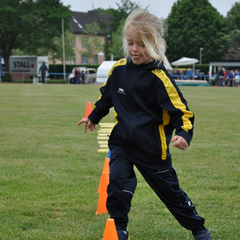 Kind in Sportkleidung balanciert auf einem Parcours aus orangefarbenen Kegeln auf einer Wiese.