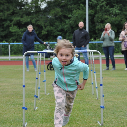 Ein Mädchen läuft fröhlich durch einen Hürdenparcours auf einem Sportfeld, während Zuschauer im Hintergrund stehen.