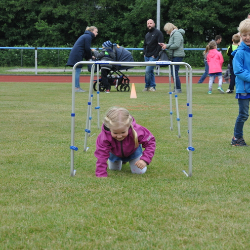 Ein kleines Mädchen kriecht unter einem Hindernis auf einem Rasenplatz, umgeben von anderen Kindern und Erwachsenen.