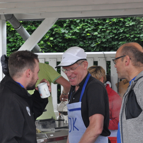Vier Personen unter einem Pavillon stehen in einem Gespräch, während im Hintergrund eine Kochstelle sichtbar ist.