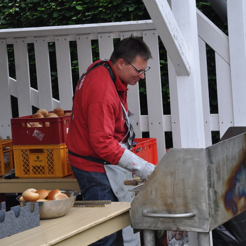 Mann in roter Jacke grillt an einem Metallgrill, während daneben Brötchen in einer Schüssel liegen.