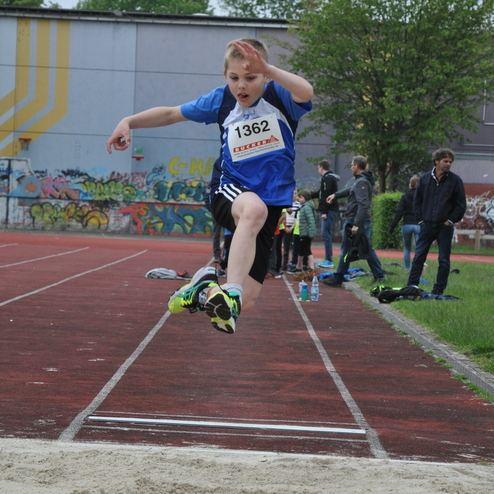 Junge springt während eines Wettkampfs in die Sandgrube eines Weitsprungbereichs. Zuschauer im Hintergrund.