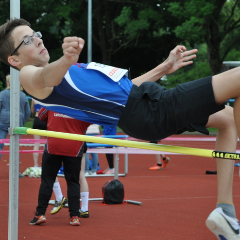 Junger Athlet springt über die Hochsprunganlage auf einem Leichtathletikplatz, im Hintergrund weitere Sportler.
