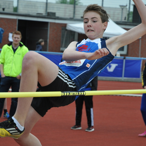 Schüler springt beim Hochsprung über eine gelbe Stange, während Zuschauer im Hintergrund auf einer Laufbahn stehen.