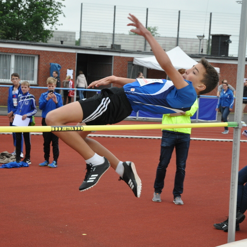 Junge springt über eine hohe Stange beim Hochsprungwettbewerb, Zuschauer beobachten am Rand des Sportplatzes.