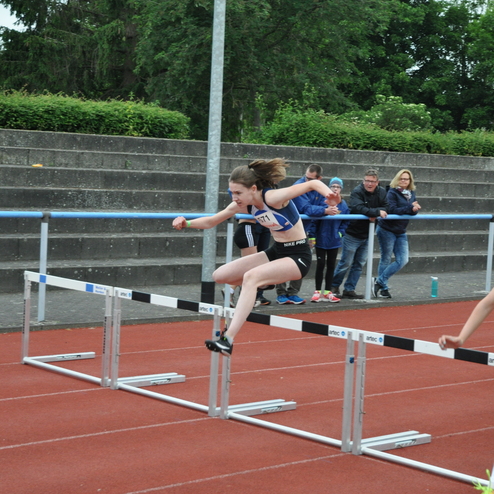 Eine Athletin springt über eine Hürde auf einer Laufbahn, Zuschauer sind im Hintergrund sichtbar.