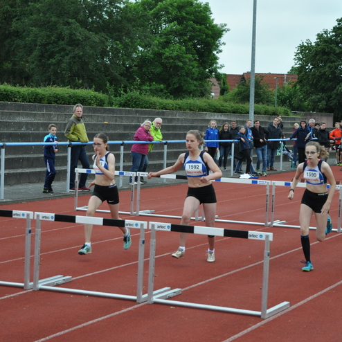 Drei junge Athletinnen überwinden Hürden auf einer roten Laufbahn, umgeben von Zuschauern auf Tribünen im Hintergrund.