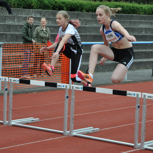 Zwei Athletinnen überwinden beim Hürdenlauf auf einer roten Laufbahn eine Hürde, Zuschauer stehen im Hintergrund.