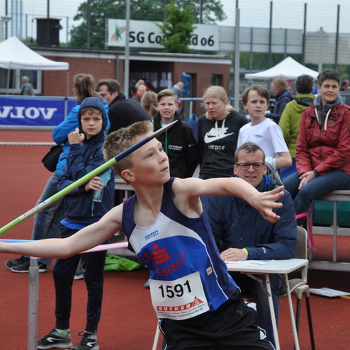 Ein athletischer Junge wirft einen Speer, während Zuschauer auf einer Sportveranstaltung im Hintergrund zuschauen.