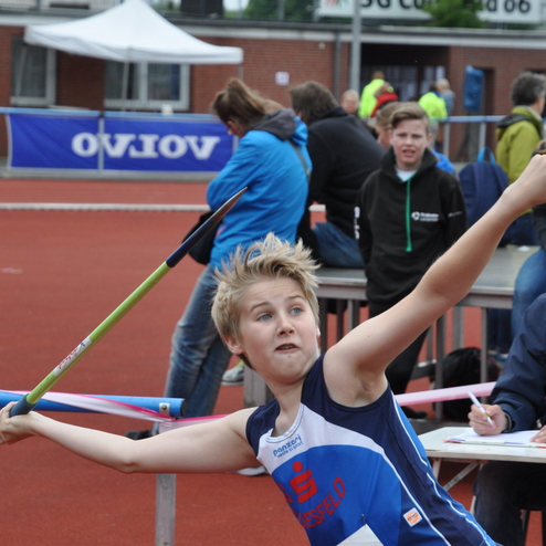 Ein Jugendlicher wirft einen Speer auf einem Sportplatz, umgeben von Zuschauern und Bewertenden.