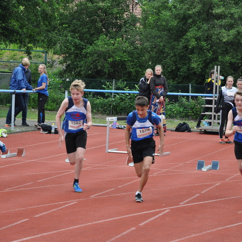 Vier Schüler sprinten über eine rote Laufbahn bei einem Sportereignis, während Zuschauer im Hintergrund zuschauen.