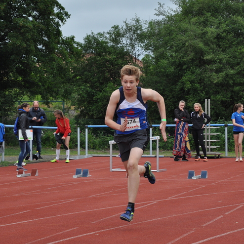Junger Athlet sprintet auf einer roten Laufbahn, umgeben von Zuschauern und anderen Sportlern.