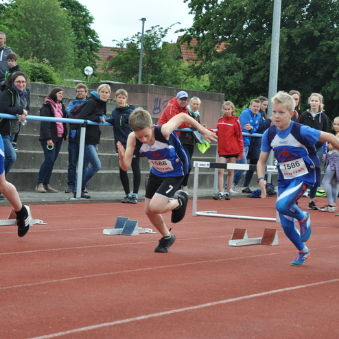 Vier junge Leichtathleten starten beim Sprint auf einer roten Tartanbahn, während Zuschauer im Hintergrund zuschauen.