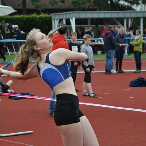 Mädchen in sportlicher Kleidung wirft einen Speer auf einem Leichtathletikfeld, Zuschauer im Hintergrund.