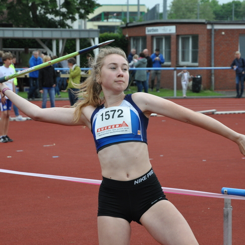 Junge Athletin wirft einen Speer auf einer roten Leichtathletikanlage, Zuschauer im Hintergrund.