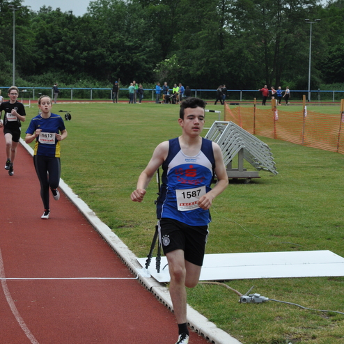 Junger Athlet in blauer Sportkleidung läuft auf einer Rennbahn, während andere Teilnehmer im Hintergrund joggen.