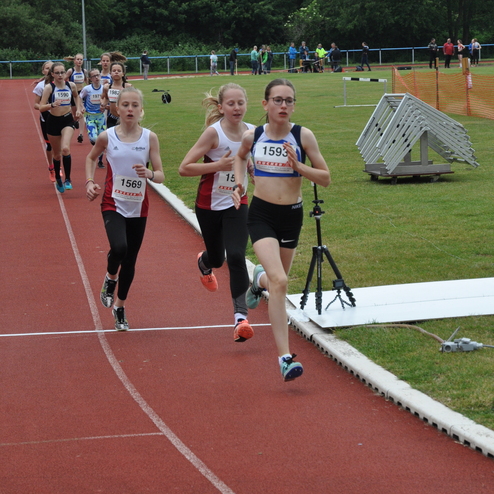 Vier junge Athletinnen laufen auf einer roten Laufbahn während eines Wettkampfs im Freien. Zuschauer stehen am Rand.